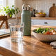 Glass of water and reusable bottle beside a meal and closed notebook on a kitchen table